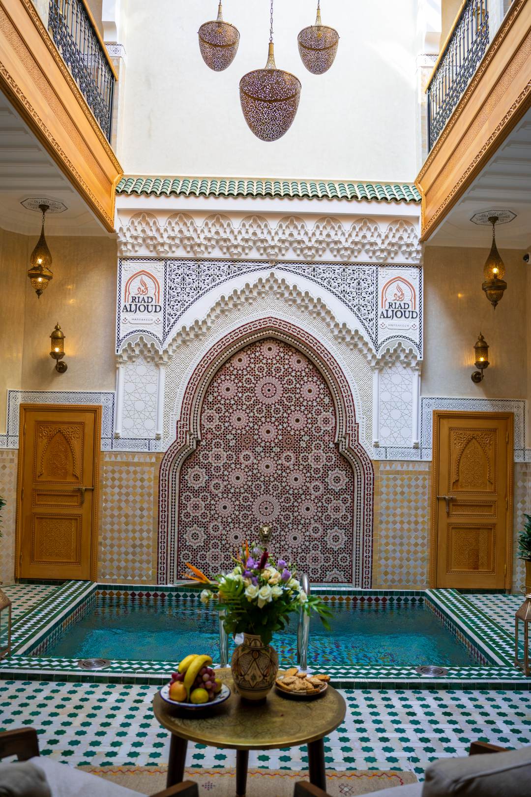 Patio du riad : bassin en zellige devant une arche de mosaïque, lanternes et galeries